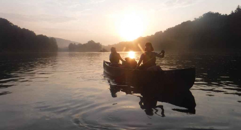 Two people paddle a canoe on calm water. The sun is low in the sky behind them. The water is framed by trees. 